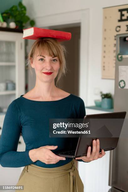 portrait of smiling woman standing with laptop in the kitchen balancing book on her head - top of head stock pictures, royalty-free photos & images