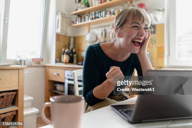 portrait of woman sitting in the kitchen with laptop crying for joy - cheering stock-fotos und bilder