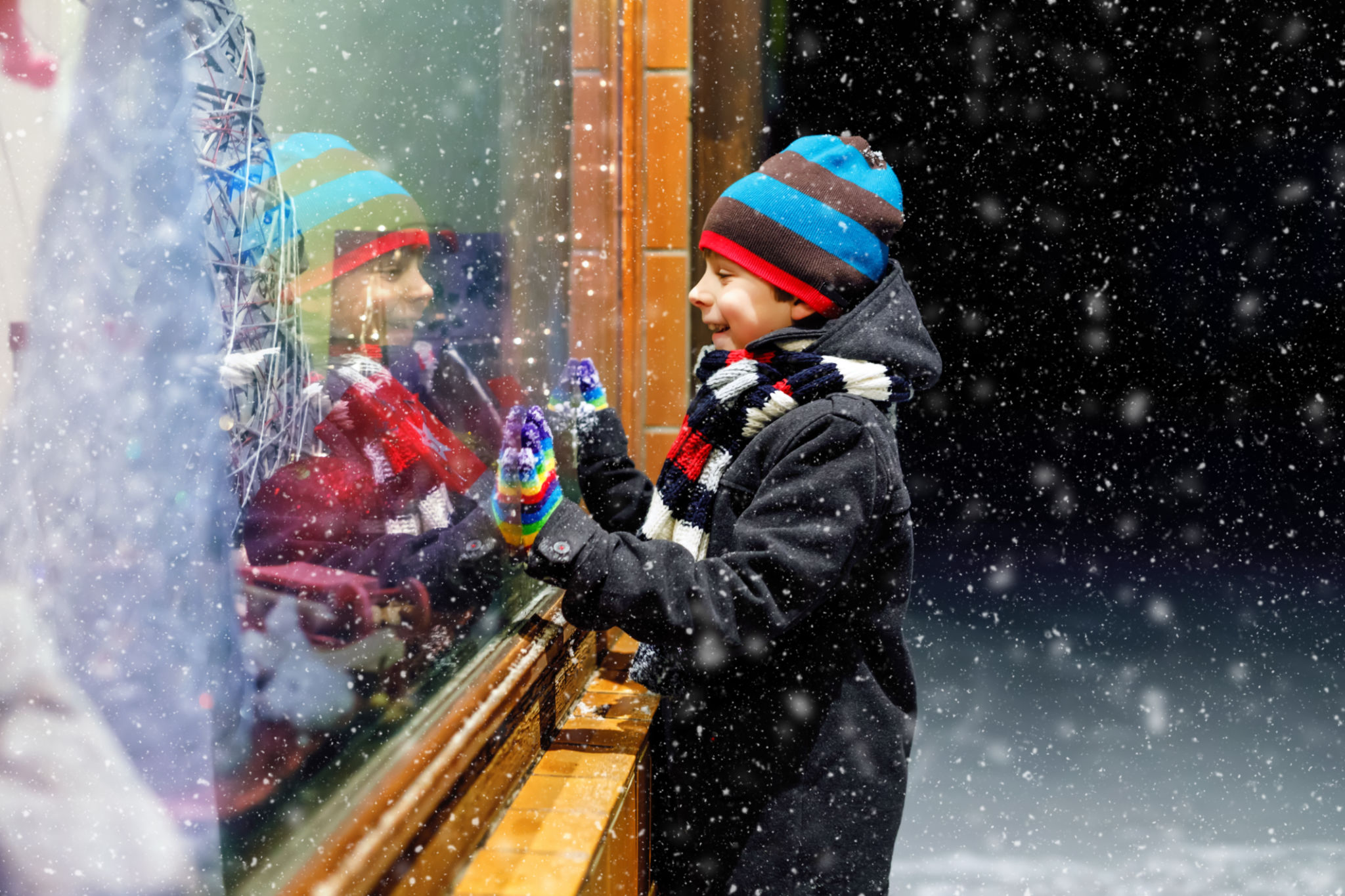 Cute little healthy school kid boy on Christmas market. Funny happy child in fashion winter clothes making window shopping decorated with gifts, xmas tree. Snow falling down, snowfall Cute little healthy school kid boy on Christmas market. Funny happy child in fashion winter clothes making window shopping decorated with gifts, xmas tree. Snow falling down, snowfall