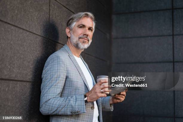 fashionable mature businessman leaning at a wall with newspaper and takeaway coffee - casual menswear stock pictures, royalty-free photos & images