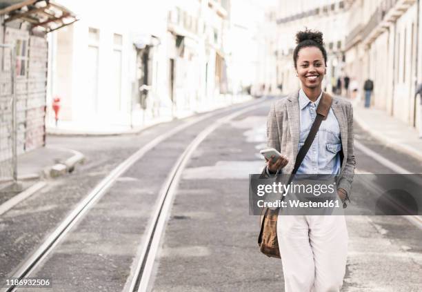 smiling young woman with mobile phone in the city on the go, lissabon, portugal - borsa a tracolla foto e immagini stock