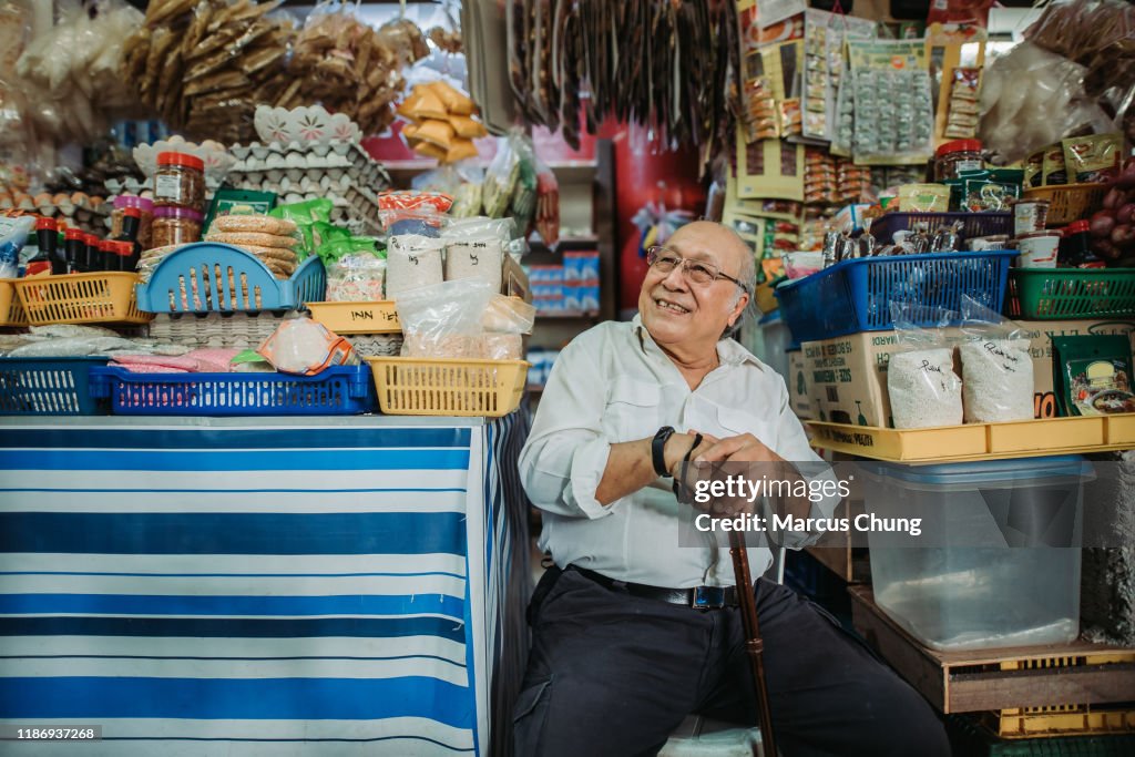 Asiático propietario de la tienda malayo de la tercera edad sentado en su tienda con su bastón para caminar, sentado y sonriendo mientras espera a que su cliente venga