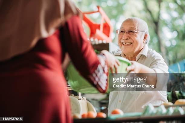 asiatische malay senior mann gemüse stall besitzer erhalten korb von gemüse von seinem kunden für die preisberechnung - malay culture stock-fotos und bilder