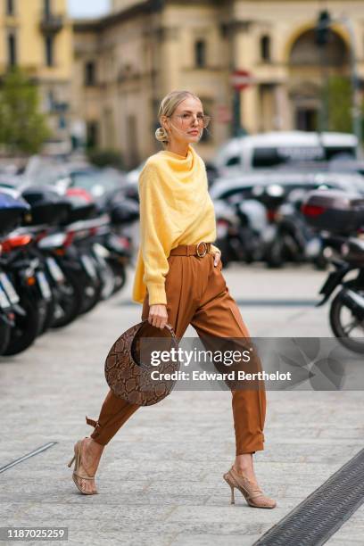 Leonie Hanne wears earrings, a fluffy yellow sweater with a scarf, rust-color pants, beige fishnet heeled pumps, a brown python pattern round...