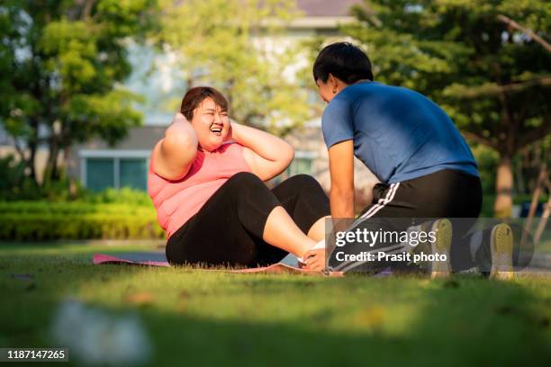 two asian trainer man and overweight woman exercising sit up together in public park in village, laughing and conversation in morning during sunlight. fat women take care of health and want to lose weight concept. - fat loss stock-fotos und bilder