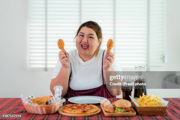 asian overweight woman holding and eating fried chicken with many junk food on the table, unhealthy nutrition and overeating. she looking happy to eat junk food. concept of eating unhealthy lifestyle. - asian eating hotdog stock-fotos und bilder