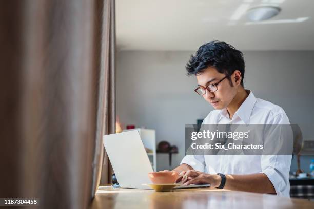 jeune homme asiatique d'affaires travaillant avec l'ordinateur portatif tout en s'asseyant dans le café de café - indonesia photos et images de collection