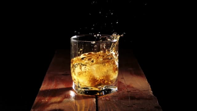 Luxury whiskey. Ice cubes fall with splashes into a glass with golden whisky on the brown wooden table against black background. Scotch in tumbler. Bourbon in highball.