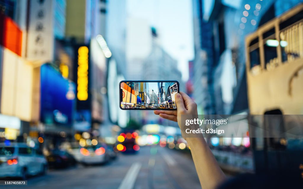 Woman's hand taking photo with smartphone in busy downtown district against urban skyscrapers with multi-coloured neon signs and city traffic