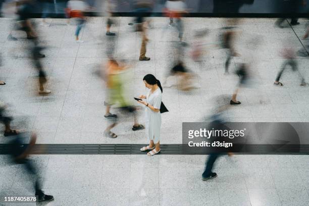 young asian woman using smartphone surrounded by commuters rushing by in subway station - crowded airport stock pictures, royalty-free photos & images