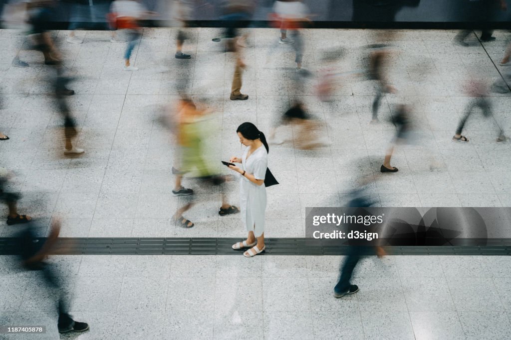 Young Asian woman using smartphone surrounded by commuters rushing by in subway station