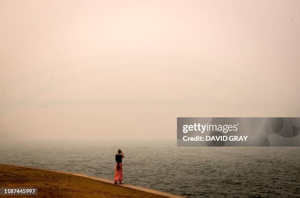 The Sydney Harbour Bridge is enveloped in haze caused by nearby bushfires as a woman takes photos on the foreshore of the Sydney Harbour in Sydney on...