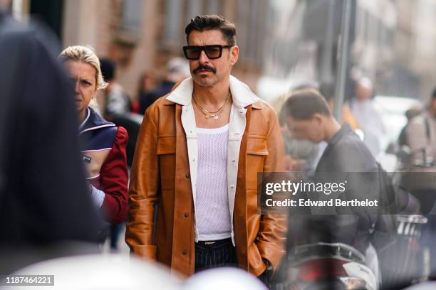 Alex Badia wears sunglasses, earrings, necklaces, a white top, a cream-color denim jacket, a tan-color leather jacket, outside the Blumarine show...