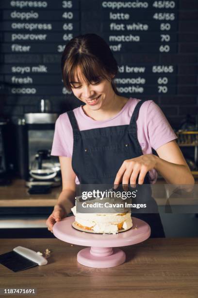 young woman in dark apron and pink t-shirt smoothing cream on a top of cake by spatula in the cafeteria. - pastelero fotografías e imágenes de stock