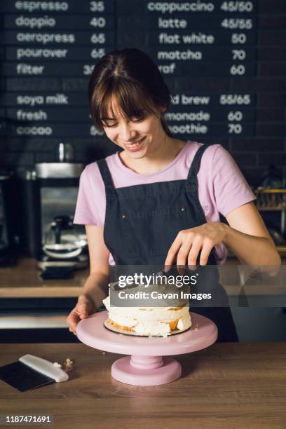 young woman in dark apron and pink t-shirt smoothing cream on a top of cake by spatula in the cafeteria. - pasteleiro imagens e fotografias de stock