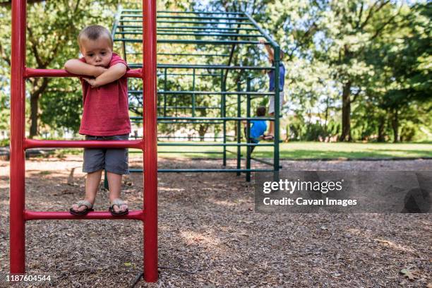 toddler boy looking sad on playground ladder with older boys playing - sad kid in kindergarten stock pictures, royalty-free photos & images