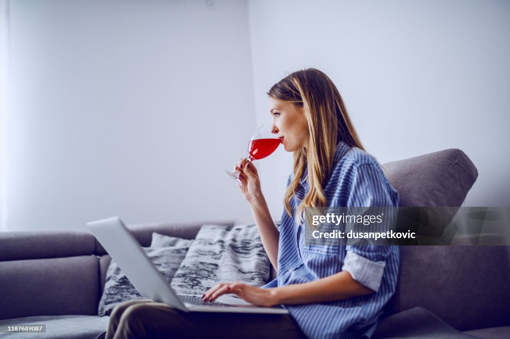 Seitenansicht der schönen kaukasischen Brünette in gestreiftem Hemd sitzen auf der Couch im Wohnzimmer, Wein trinken und mit Laptop.