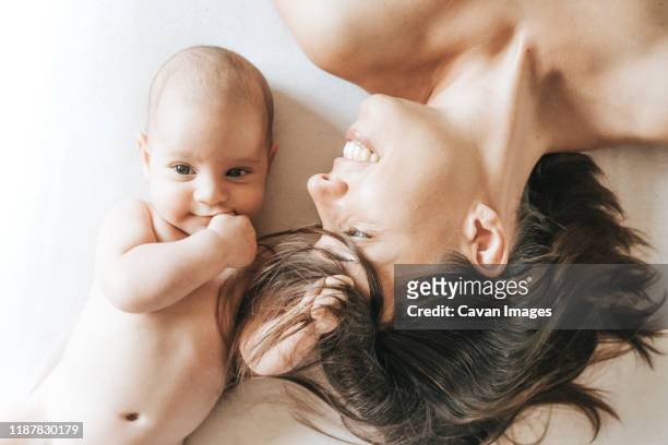 mother and newborn son stretched out in bed, the son catches the mother's hairs and she looks at him with love - baby skin fotografías e imágenes de stock