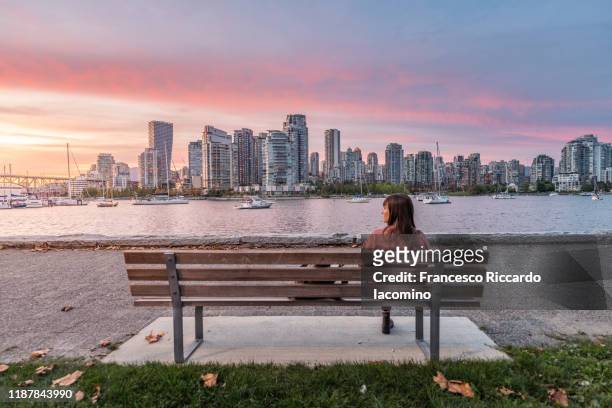woman sitting on a bench looking at vancouver skyline, sunset from from the island park walk. british columbia, canada - vancouver canada foto e immagini stock