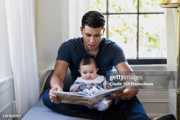 zittend bij venster, papa leest boek aan baby - op schoot stockfoto's en -beelden