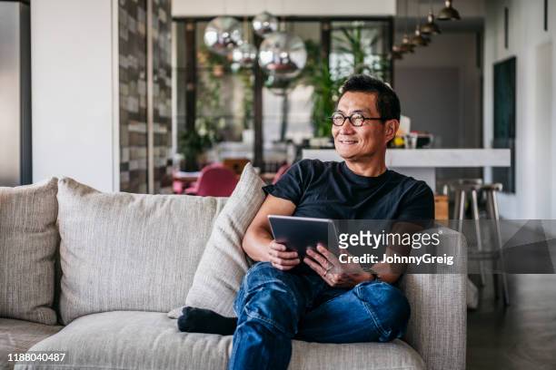 cheerful chinese man with tablet looking away and smiling - asian man sitting casual imagens e fotografias de stock