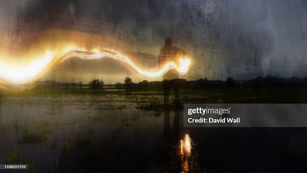 A deliberately blurred, abstract, out of focus, textured edit. Of a ghostly, eerie figure, standing in a flooded field, holding a glowing lamp with a flaming light trail behind. On a winters evening.