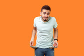 Portrait of frustrated worried brunette man turning out empty pockets. indoor studio shot isolated on orange background