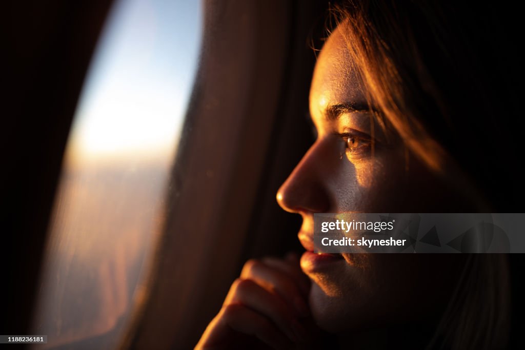 Primer plano de la mujer pensativa mirando la puesta del sol a través de la ventana del avión.