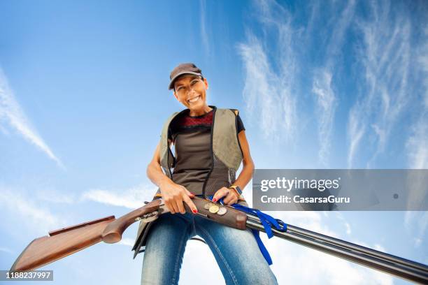 porträt von smiling mid adult woman holding shotgun against the sky - trap shooting stock-fotos und bilder