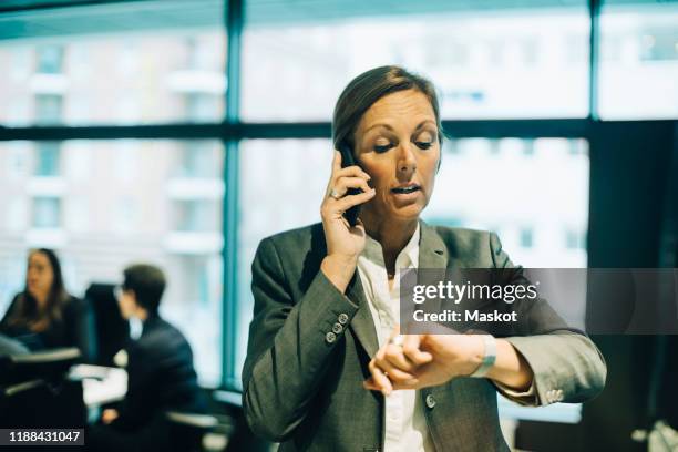 busy businesswoman talking on mobile phone while checking time on watch at office - auf die uhr sehen stock-fotos und bilder