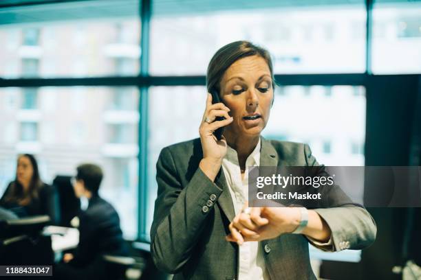 busy businesswoman talking on mobile phone while checking time on watch at office - puntualidad fotografías e imágenes de stock