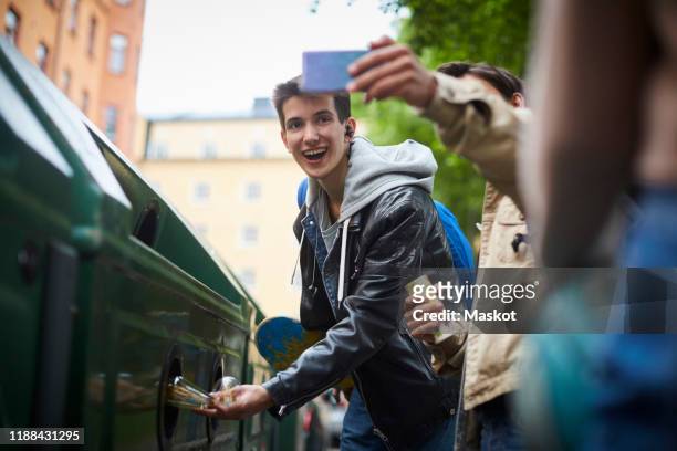 man taking selfie with teenage friend throwing waste in garbage bin at recycling station - throwing phone stock pictures, royalty-free photos & images