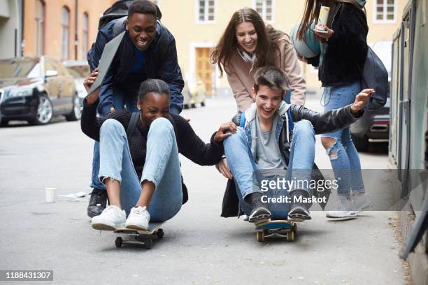 cheerful friends pushing teenagers sitting on skateboard - grupo de adolescentes imagens e fotografias de stock