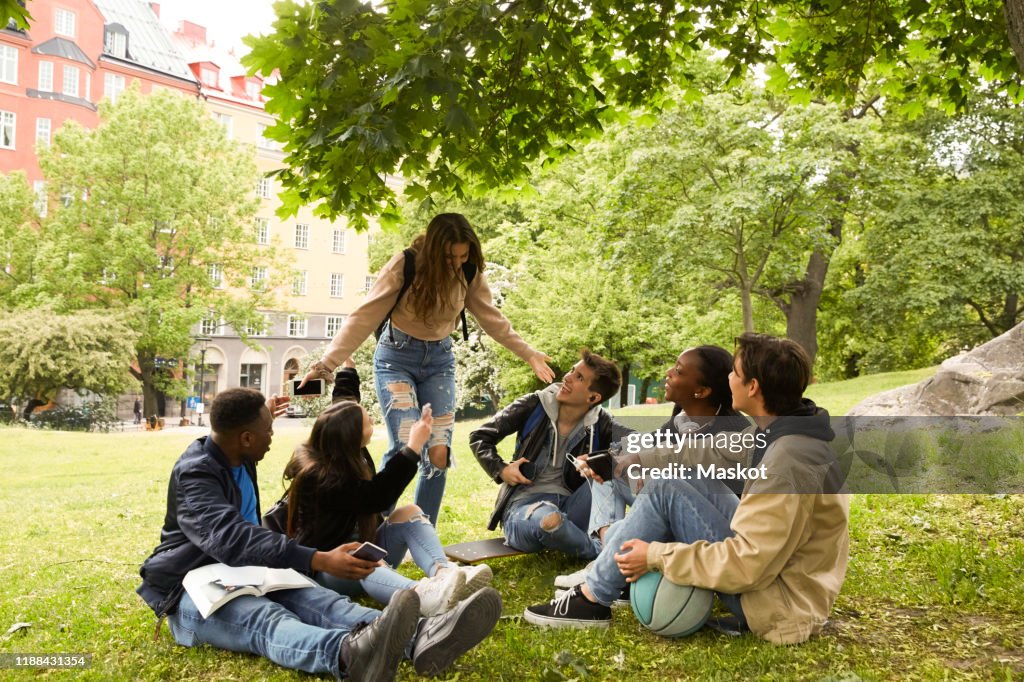 Happy teenage girl gesturing while standing by friends sitting on land at park