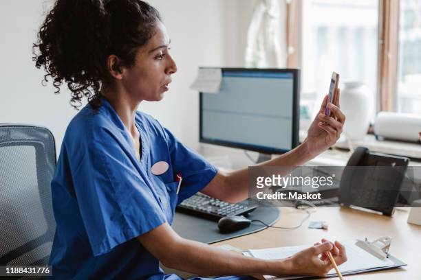 female healthcare worker talking on video conference through mobile phone in doctor's office - doctor smartphone stock pictures, royalty-free photos & images