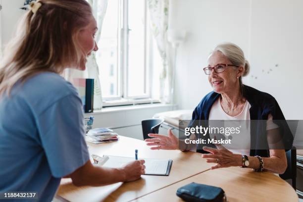 smiling doctor writing prescription while listening to senior woman in hospital - patientin stock-fotos und bilder