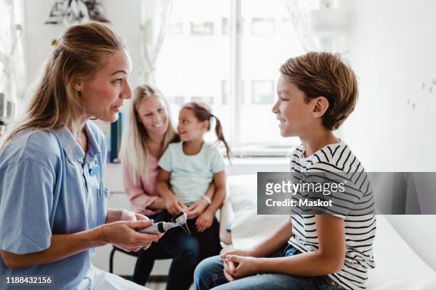 smiling female pediatrician with otoscope talking to boy while family sitting in background at medical clinic - otoscope bildbanksfoton och bilder