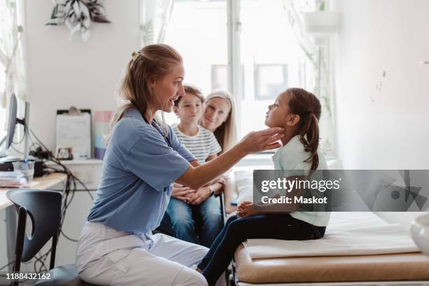 female doctor examining throat of girl while family sitting in medical room - pédiatre photos et images de collection