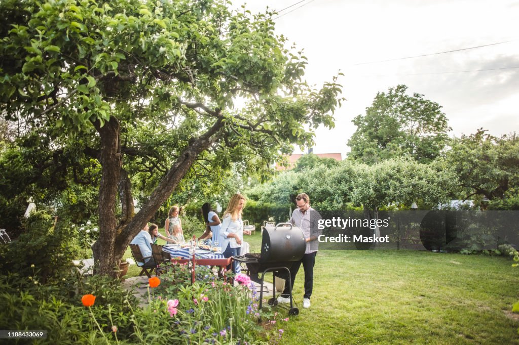Male and female friends preparing food on barbecue while family having fun in backyard