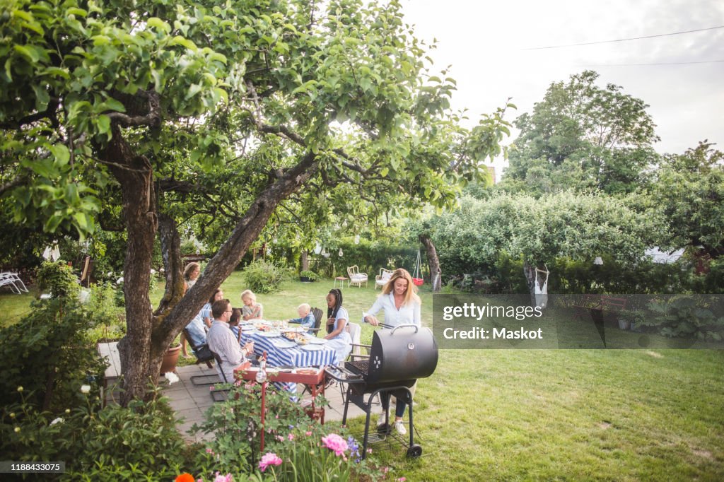 Female preparing food o barbecue while family sitting by table in backyard