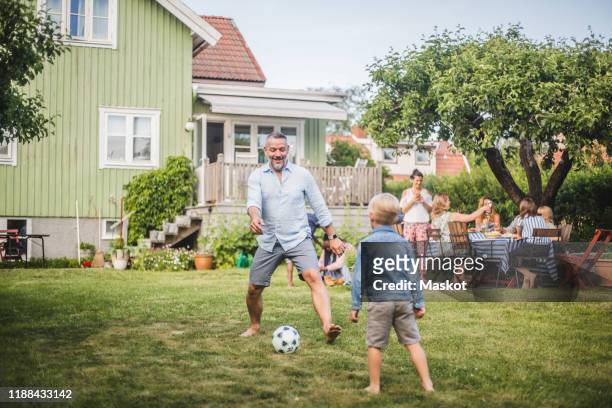 father playing football with son while friends having fun at table in backyard party - mature men friends stock pictures, royalty-free photos & images