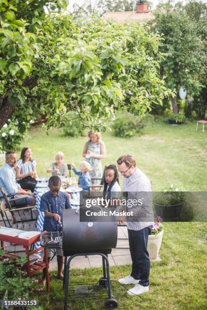 high angle view of man preparing barbecue grill with boy while family and friends sitting at table in backyard - grill party stockfoto's en -beelden