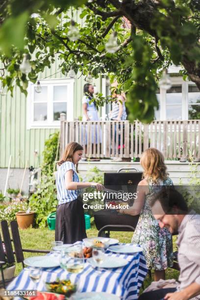 daughter and mother preparing barbecue food while smiling man sitting at table in backyard during weekend - backyard grilling stockfoto's en -beelden