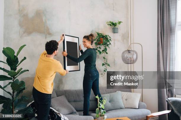 mid adult man passing picture frame to girlfriend against wall at new home - finishing touch stock-fotos und bilder