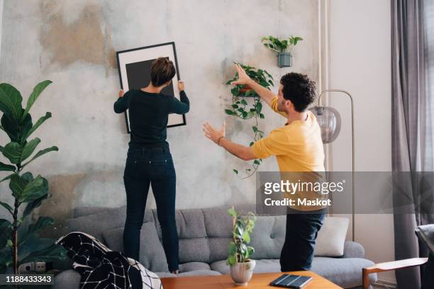 mid adult man guiding girlfriend in hanging picture frame on wall at new home - interiorismo fotografías e imágenes de stock
