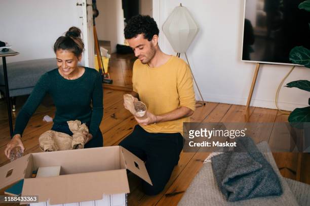 couple removing glasses from box in living room at new home - man with moving boxes authentic stockfoto's en -beelden