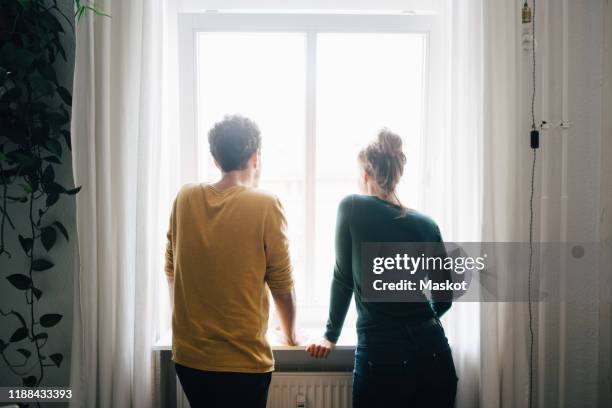 rear view of couple looking through window while standing at home - menselijke relaties stockfoto's en -beelden