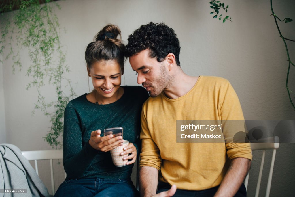 Smiling girlfriend showing smart phone to boyfriend while having coffee in living room