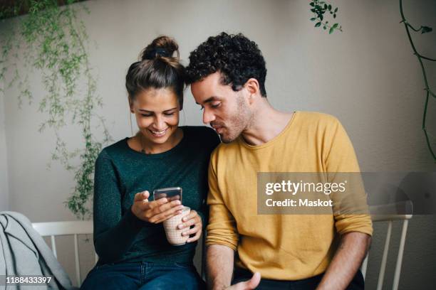 smiling girlfriend showing smart phone to boyfriend while having coffee in living room - män i 30 årsåldern bildbanksfoton och bilder