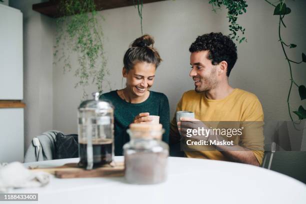smiling boyfriend and girlfriend having coffee at table in living room - coffee plunger stockfoto's en -beelden
